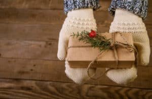 A pair of hands wearing mittens offering a gift wrapped in brown paper and decorated with string and a sprig of evergreen.