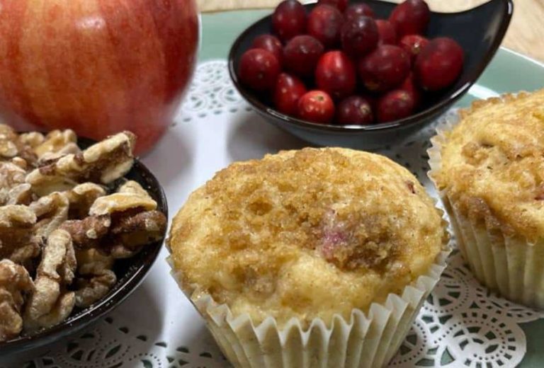 Two apple cranberry walnut muffins sit on a plate with a red apple, some walnuts and cranberries gathered around them.